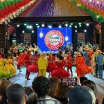 Integrantes de quadrilha junina estudantil dançando caracterizados com trajes caipiras coloridos sobre o tablado de apresentação.