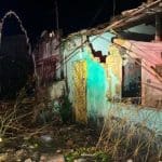 Traseira de um caminhão de carga de botijões de gás cravada na parede destruída de uma casa, com equipes dos Bombeiros e uma retroescavadeira ao redor.