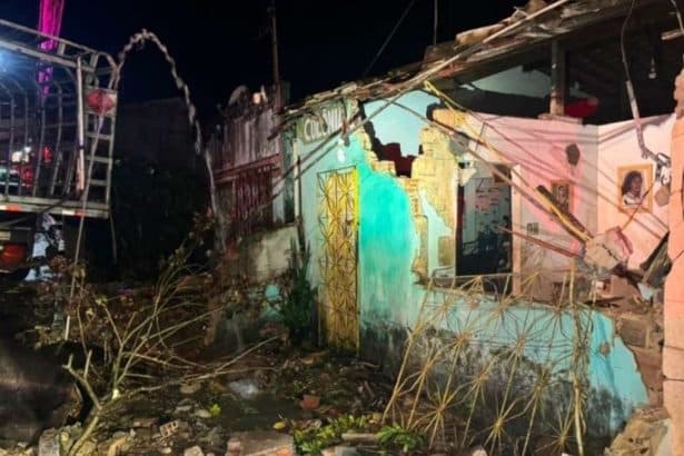 Traseira de um caminhão de carga de botijões de gás cravada na parede destruída de uma casa, com equipes dos Bombeiros e uma retroescavadeira ao redor.