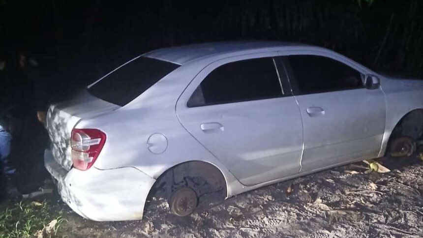 Traseira e lateral de um veículo Chevrolet Cobalt branco com marcas de poeira, estacionado em meio ao mato de uma estrada de terra na zona rural.